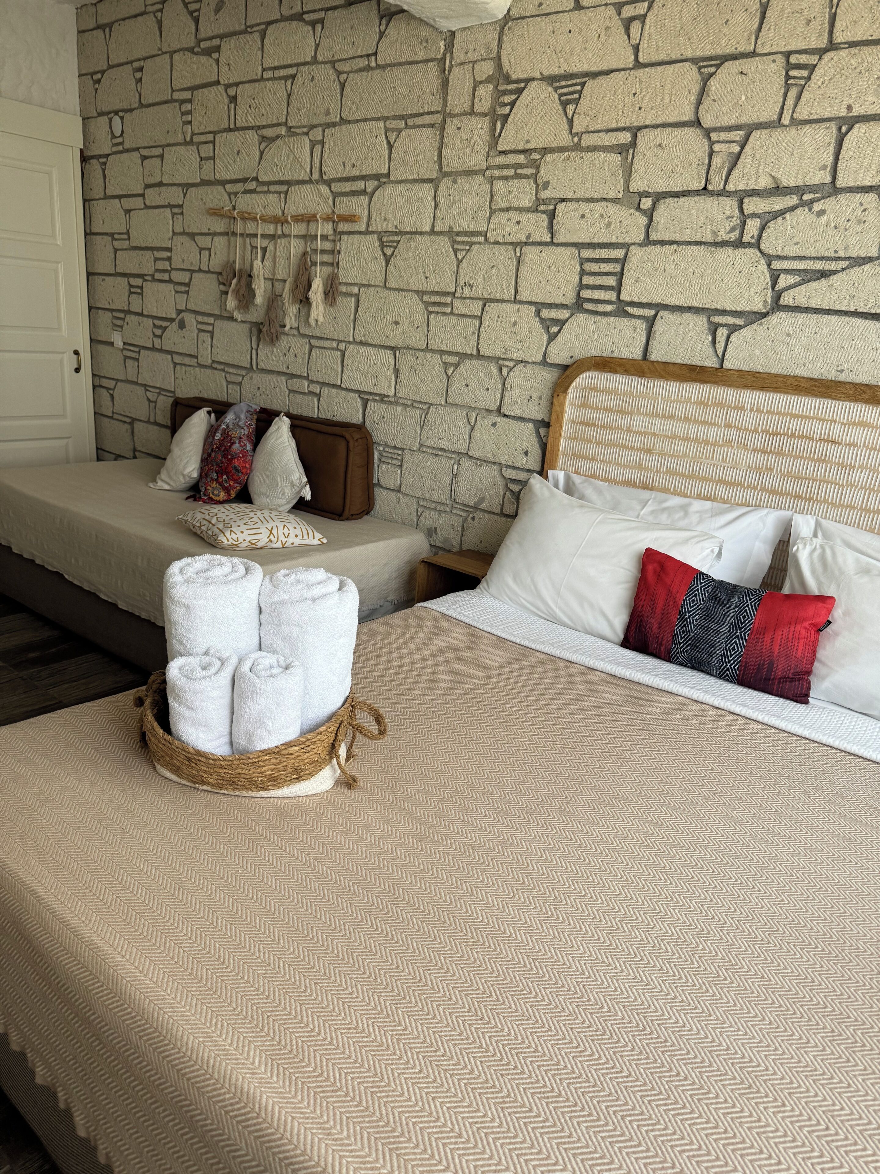 Habitación de hotel con pared de piedra, una cama con ropa de cama blanca, una mesita de noche con una cesta de toallas dobladas y un cabecero de madera.