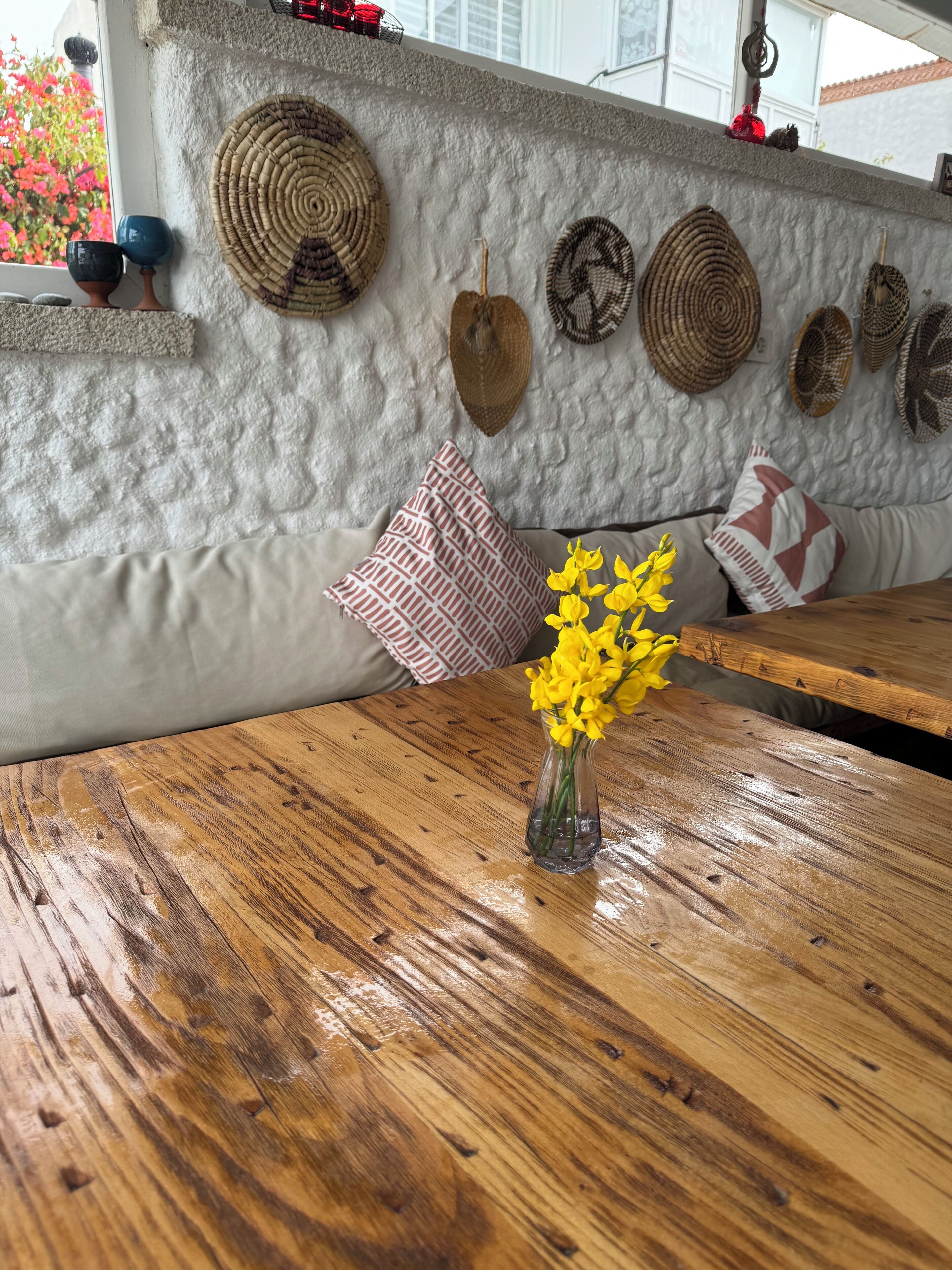 A wooden table with a vase of yellow flowers, a textured wall with woven baskets, and patterned cushions.