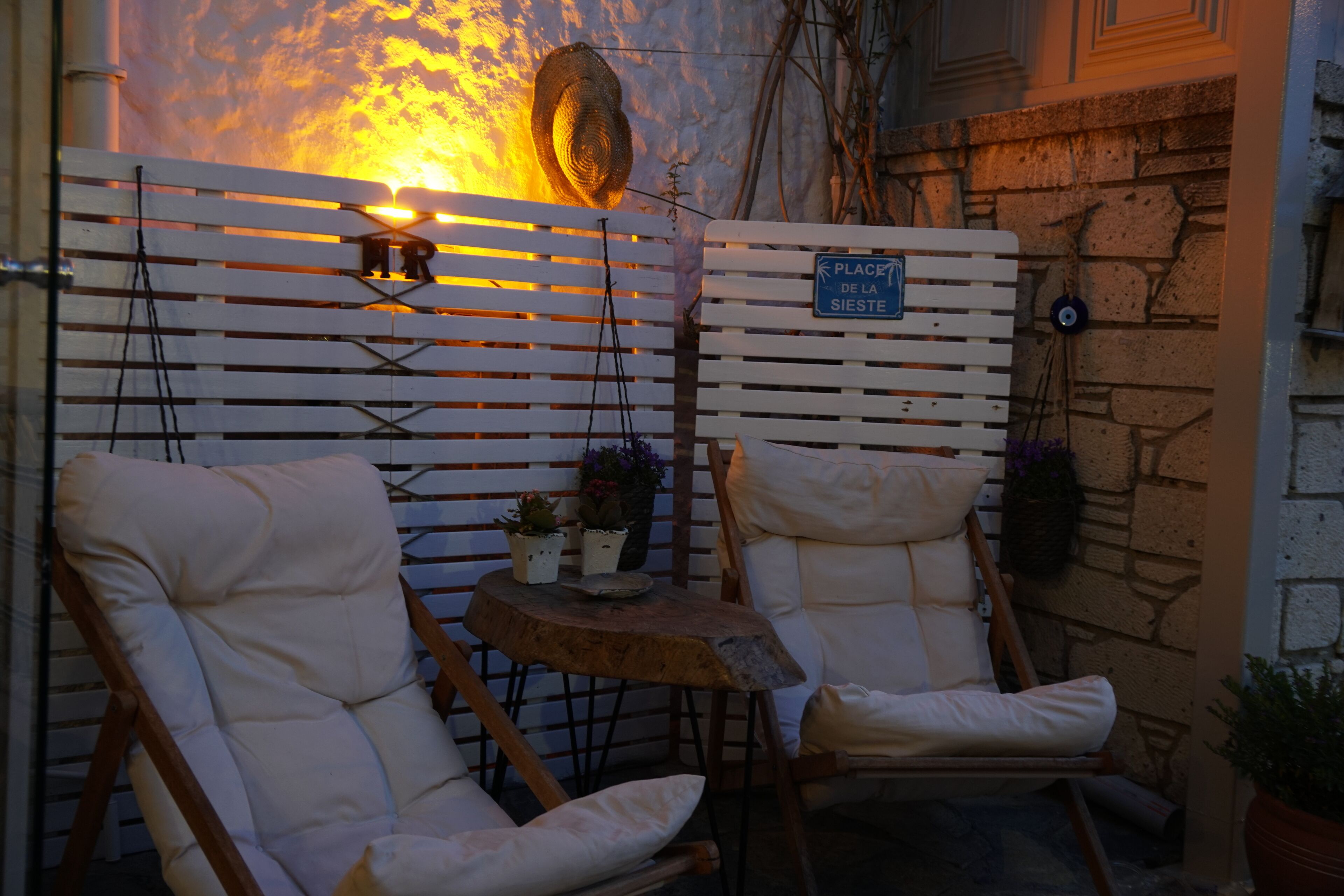 Two white cushioned chairs with a small wooden table between them, a sign reading "Place de la Siete," and a stone wall in the background.