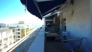 A balcony with a blue awning, metal chairs, and a view of the sea and buildings. - Vera Cruz A4 (Lido di Jesolo)