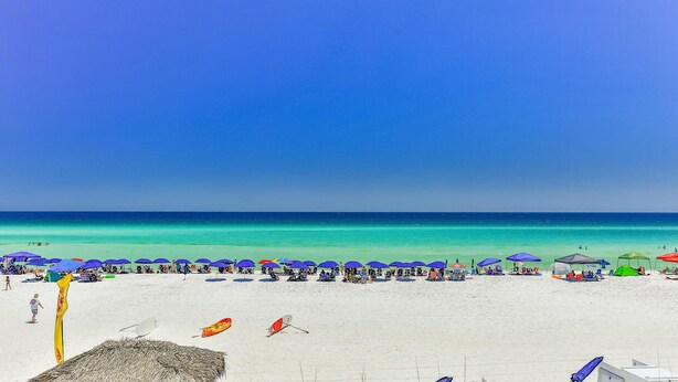 Ein Strand mit weißem Sand, klarem türkisblauem Wasser und blauem Himmel. Entlang der Küste befinden sich mehrere Sonnenschirme und Strandstühle.