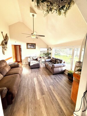 A spacious living room with a high ceiling, a ceiling fan, and a view of the outdoors through large windows. - Paradise Suites, Homer Alaska, an entire house. private hot tub (Homer)