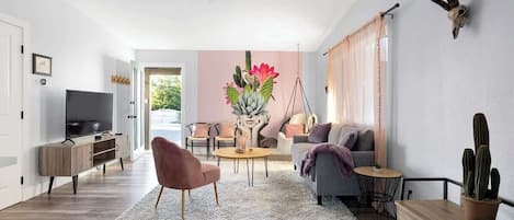 Uma sala de estar moderna com uma televisão de tela plana, uma poltrona rosa, um sofá cinza, uma mesa de centro redonda e um cacto sobre uma mesa de madeira.