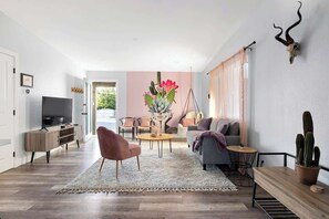 A modern living room with a flat-screen TV, a pink chair, a grey sofa, a round coffee table, and a cactus on a wooden table.