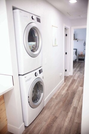 A laundry room with two stacked washing machines, a wooden floor, and a door leading to another room. - 7 Mins to Joshua Tree Park, Hot Tub · Sunset Jewel (Joshua Tree)