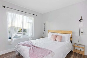 A bedroom with a large bed, a window with sheer curtains, a bedside table, and a wall-mounted lamp. - 7 Mins to Joshua Tree Park, Hot Tub · Sunset Jewel (Joshua Tree)