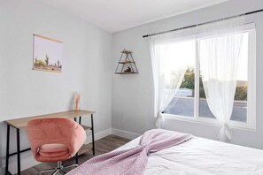A bedroom with a bed, a desk, a chair, a window with curtains, and a wall-mounted shelf.