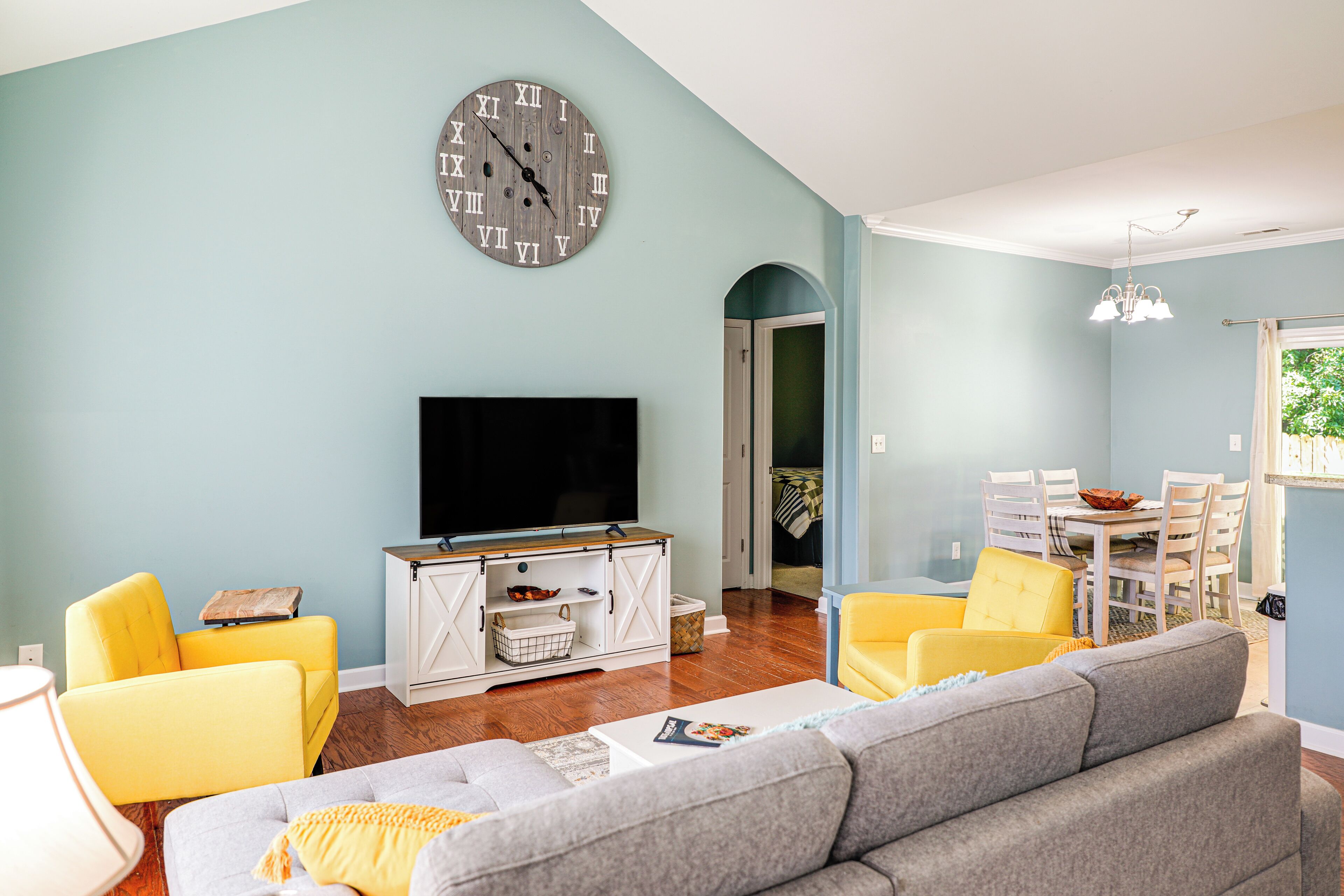 Una sala de estar con una televisión de pantalla plana, un sofá gris, sillas amarillas y una zona de comedor con mesa y sillas.