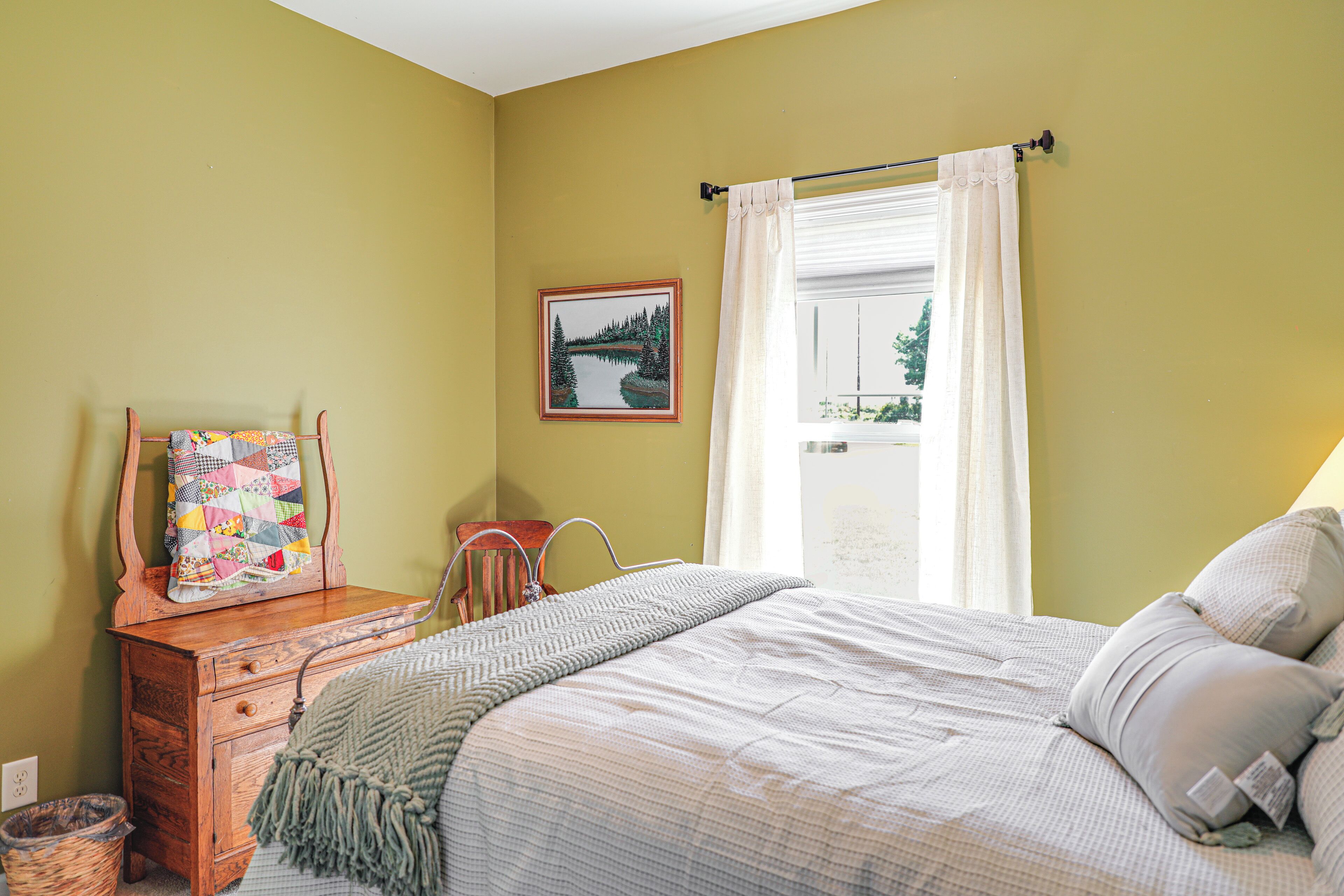 A bedroom with a bed, a wooden dresser, a chair, a window with curtains, and a framed picture on the wall.