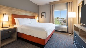A hotel room with a large bed, a nightstand with a lamp, a dresser, and a window with a view of the outdoors. - Candlewood Suites Oxford - Anniston By IHG (Oxford)
