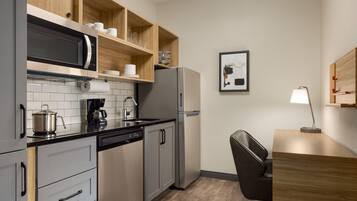A modern kitchen with a microwave, coffee maker, and refrigerator. There is a wooden shelf above the sink with cups and glasses. A chair and desk are visible in the corner.