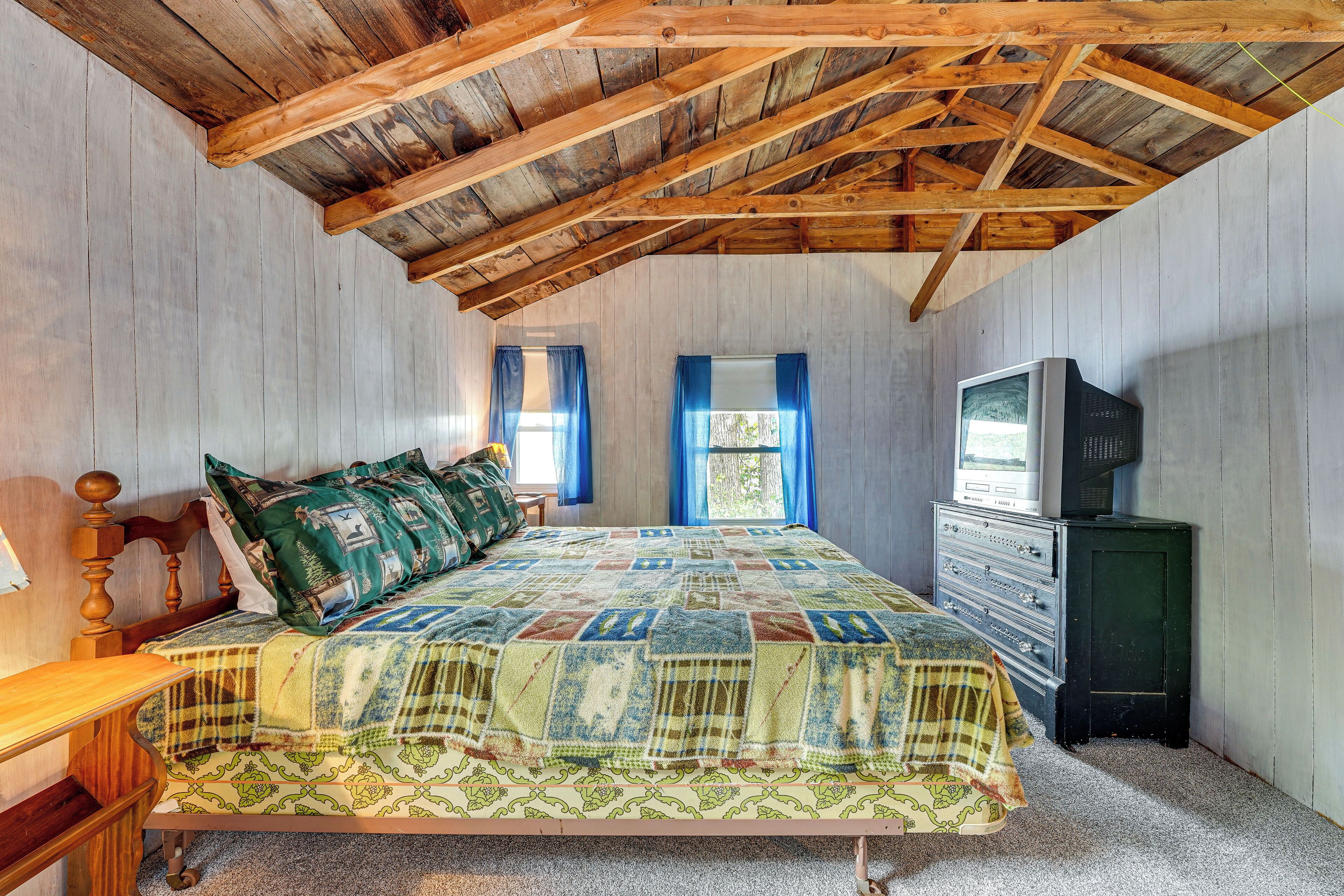 A bedroom with a wooden ceiling, a bed with a quilt, a dresser, and a television.