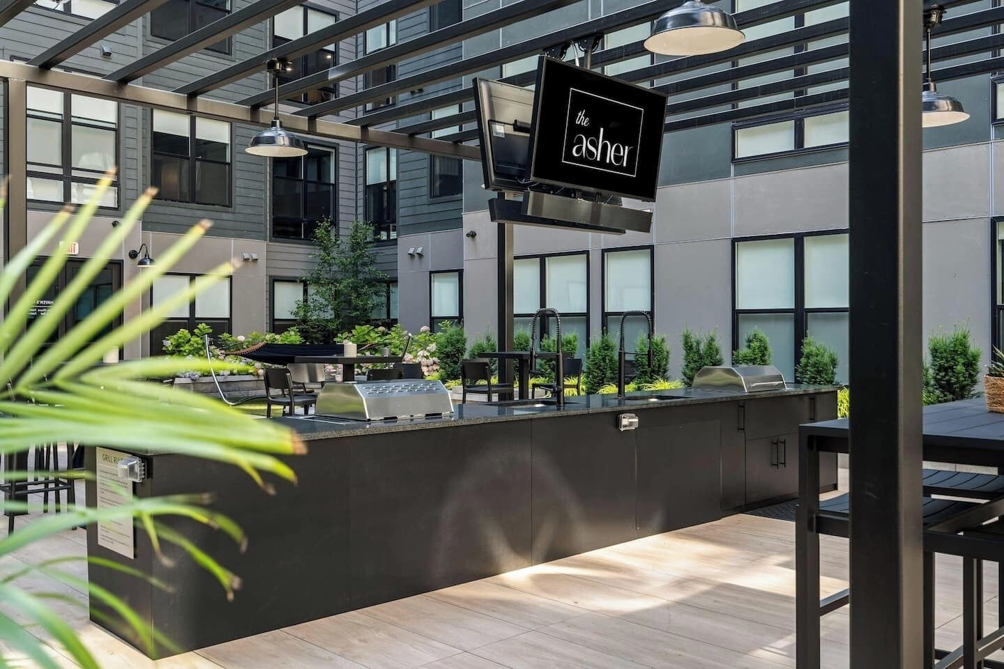 An outdoor seating area with a grill station, tables, and chairs, surrounded by greenery and modern architecture.