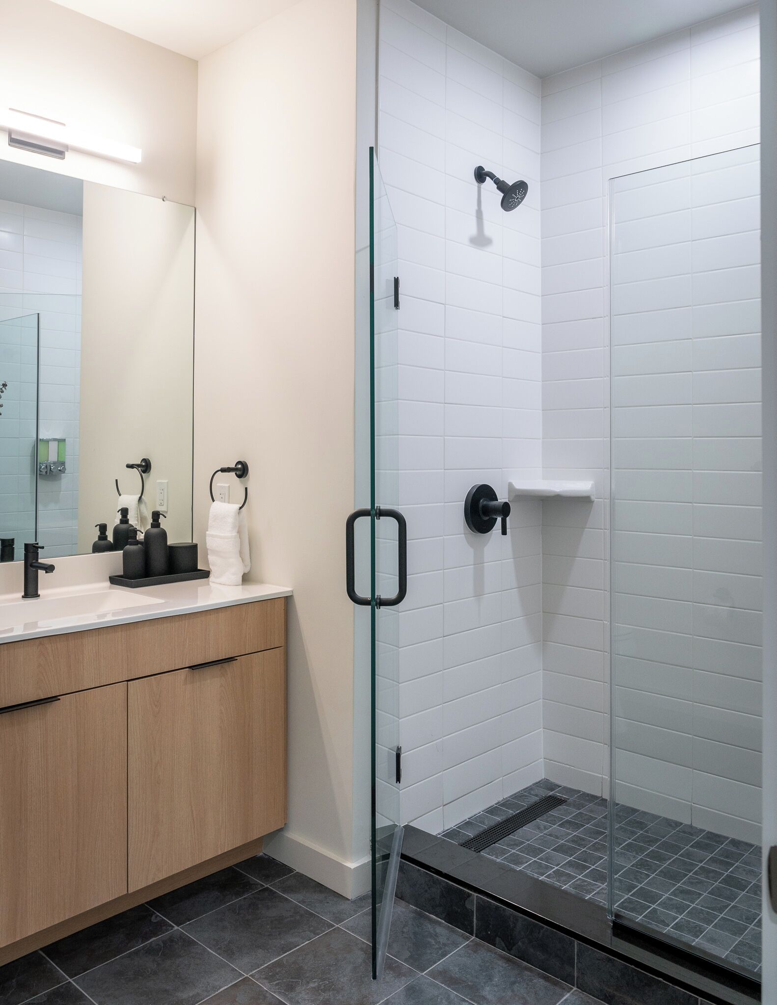 A modern bathroom with a glass shower enclosure, a white tiled wall, a wooden vanity, and a mirror.