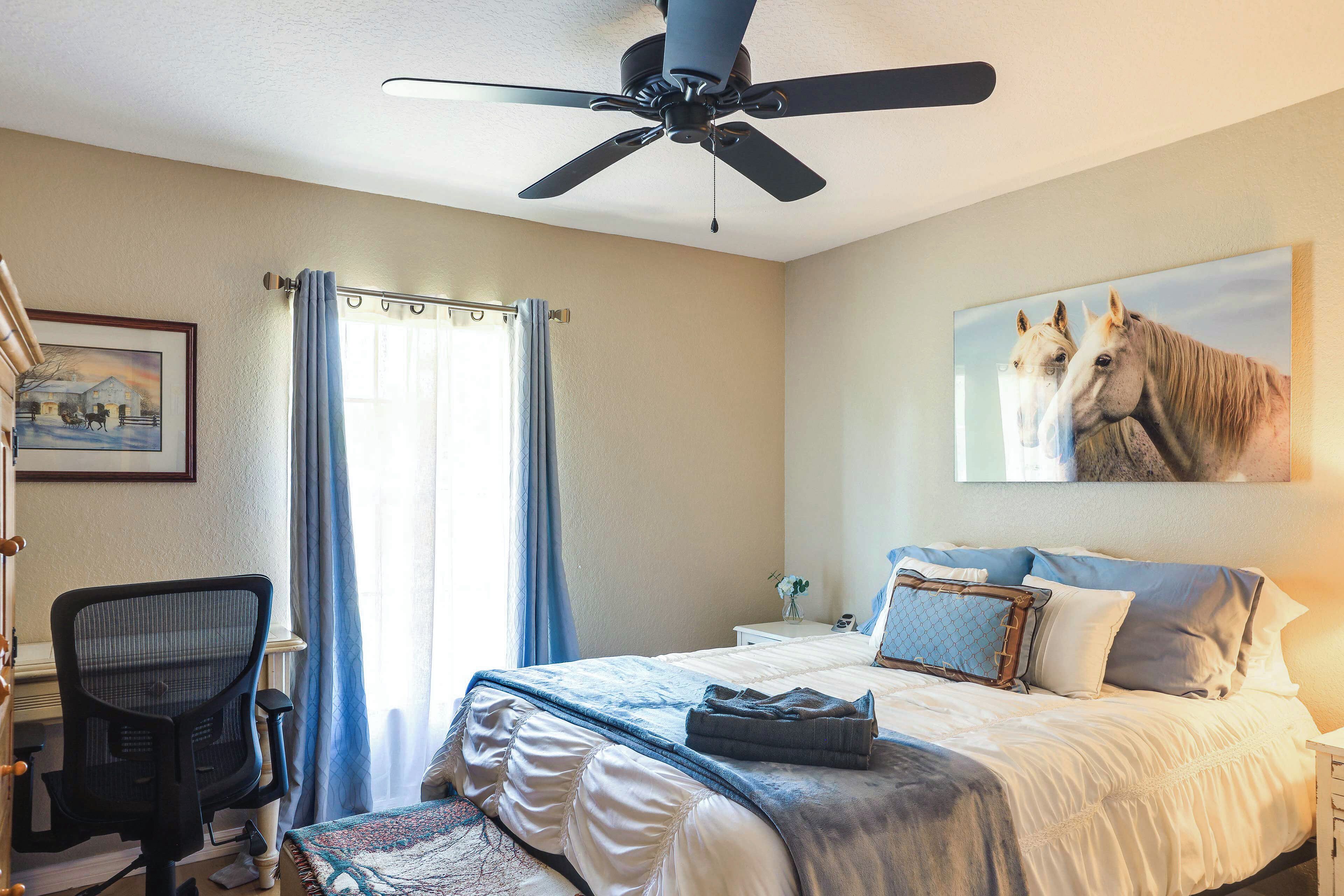 A bedroom with a bed, a desk, a chair, a ceiling fan, and a painting of horses.