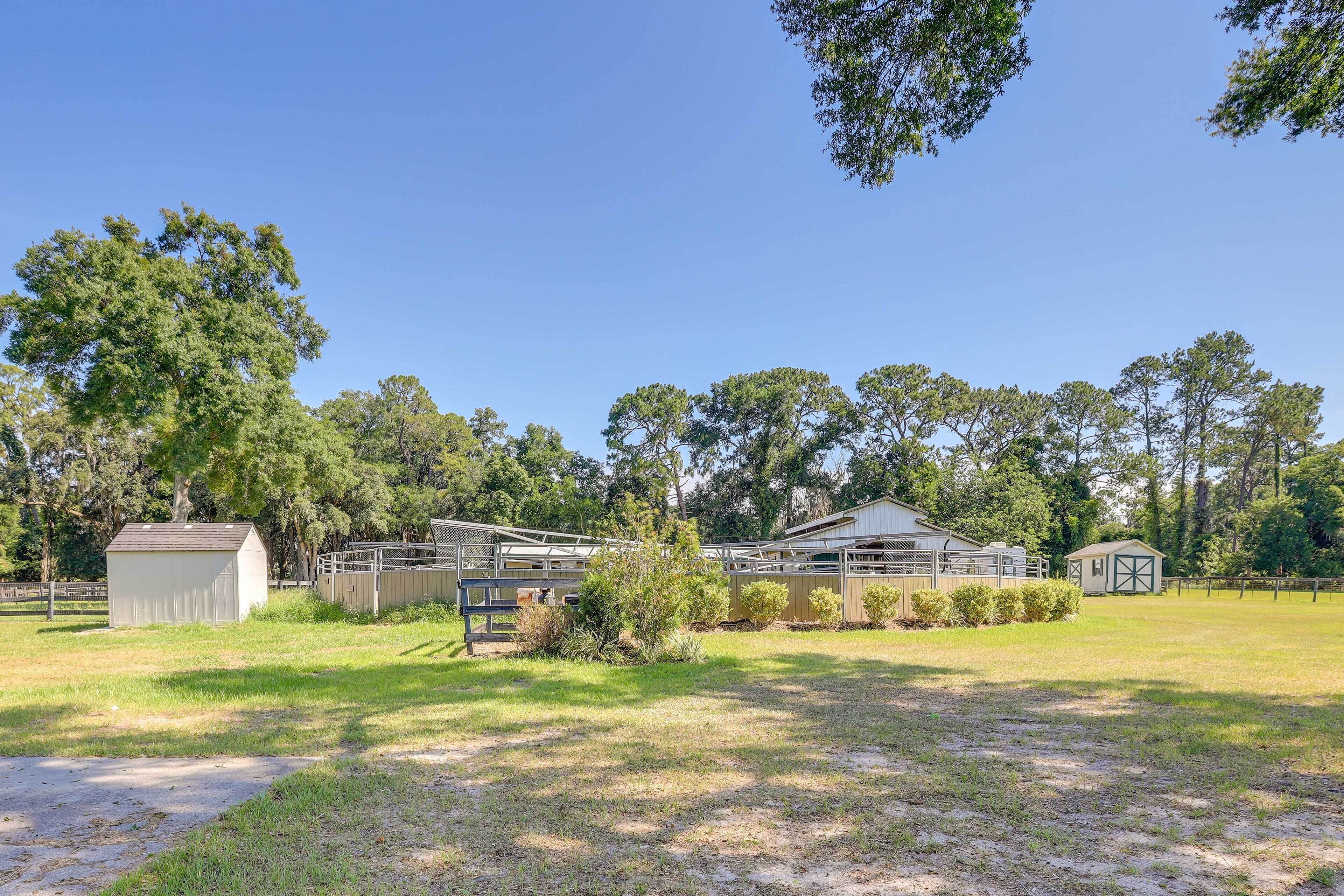 A stable with a fenced area, a small shed, and a large open field with trees in the background.