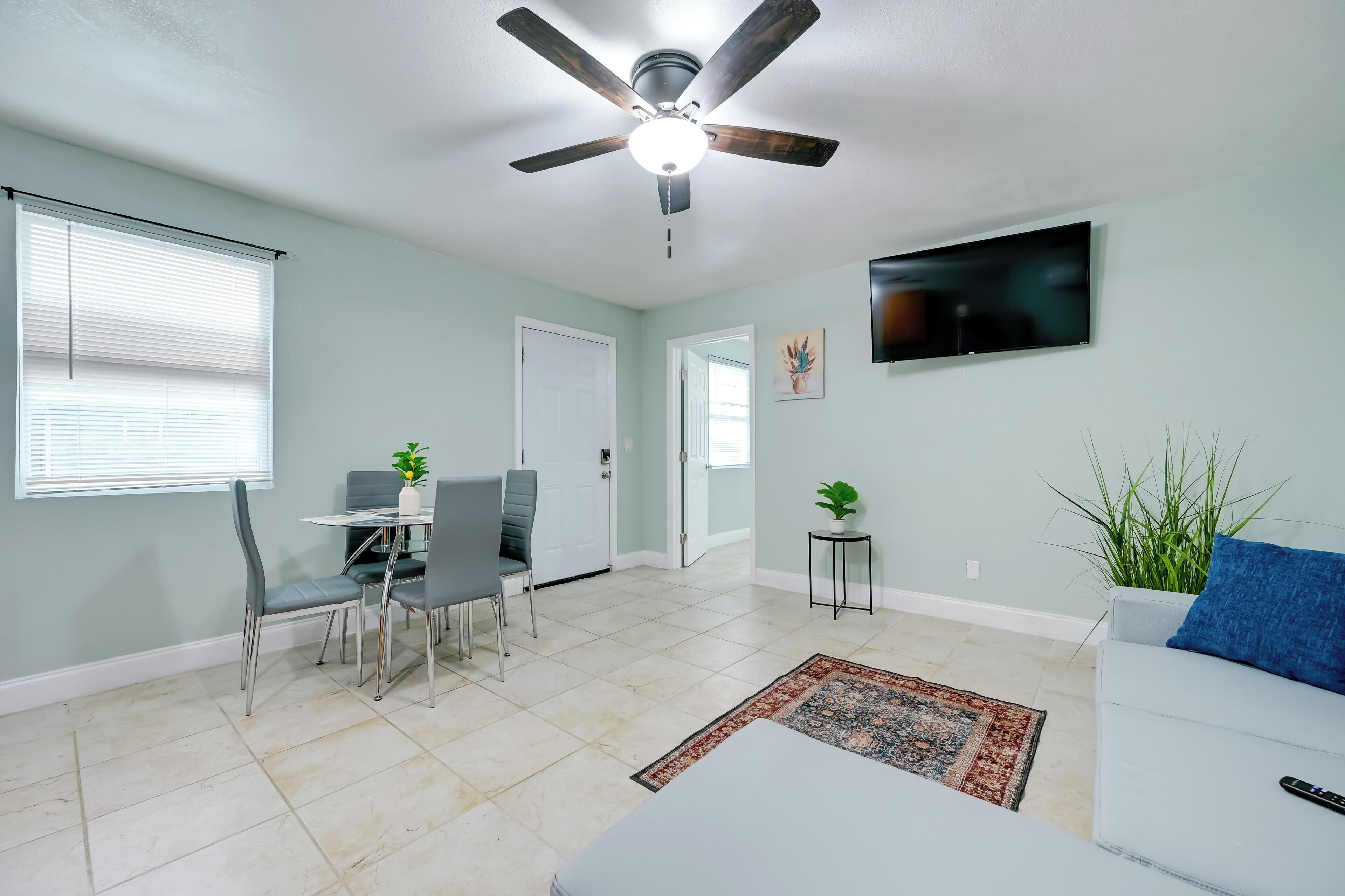 A living room with a ceiling fan, a dining area with chairs and a table, a TV mounted on the wall, and a couch with a remote on it.