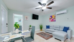 A living room with a ceiling fan, a grey sofa, a TV mounted on the wall, and a dining area with a glass table and chairs. - Modern Tampa Apartment ~ 3 Mi to Busch Gardens! (Tampa)