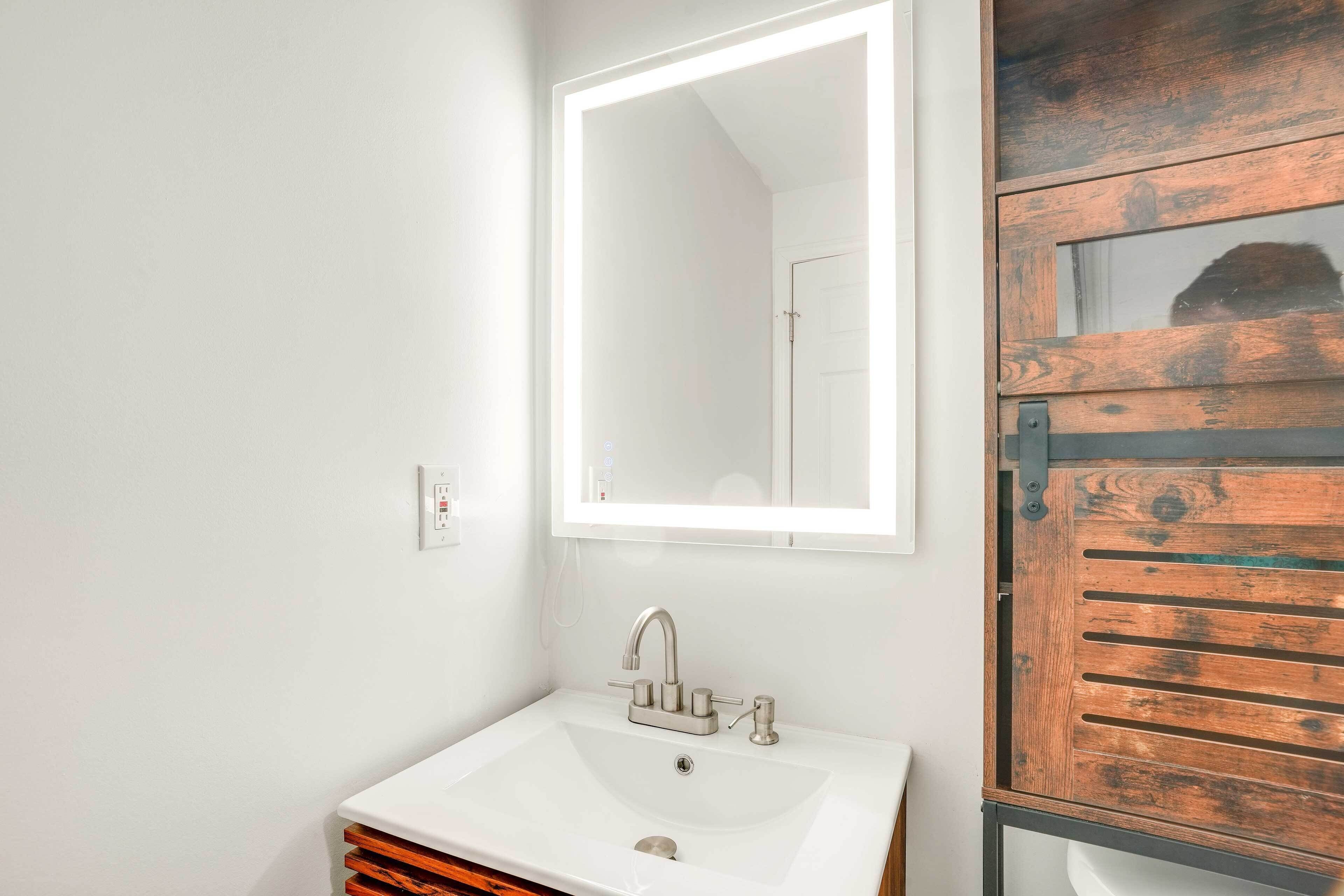 Un baño con un lavamanos blanco, un espejo con luz y una puerta de madera.