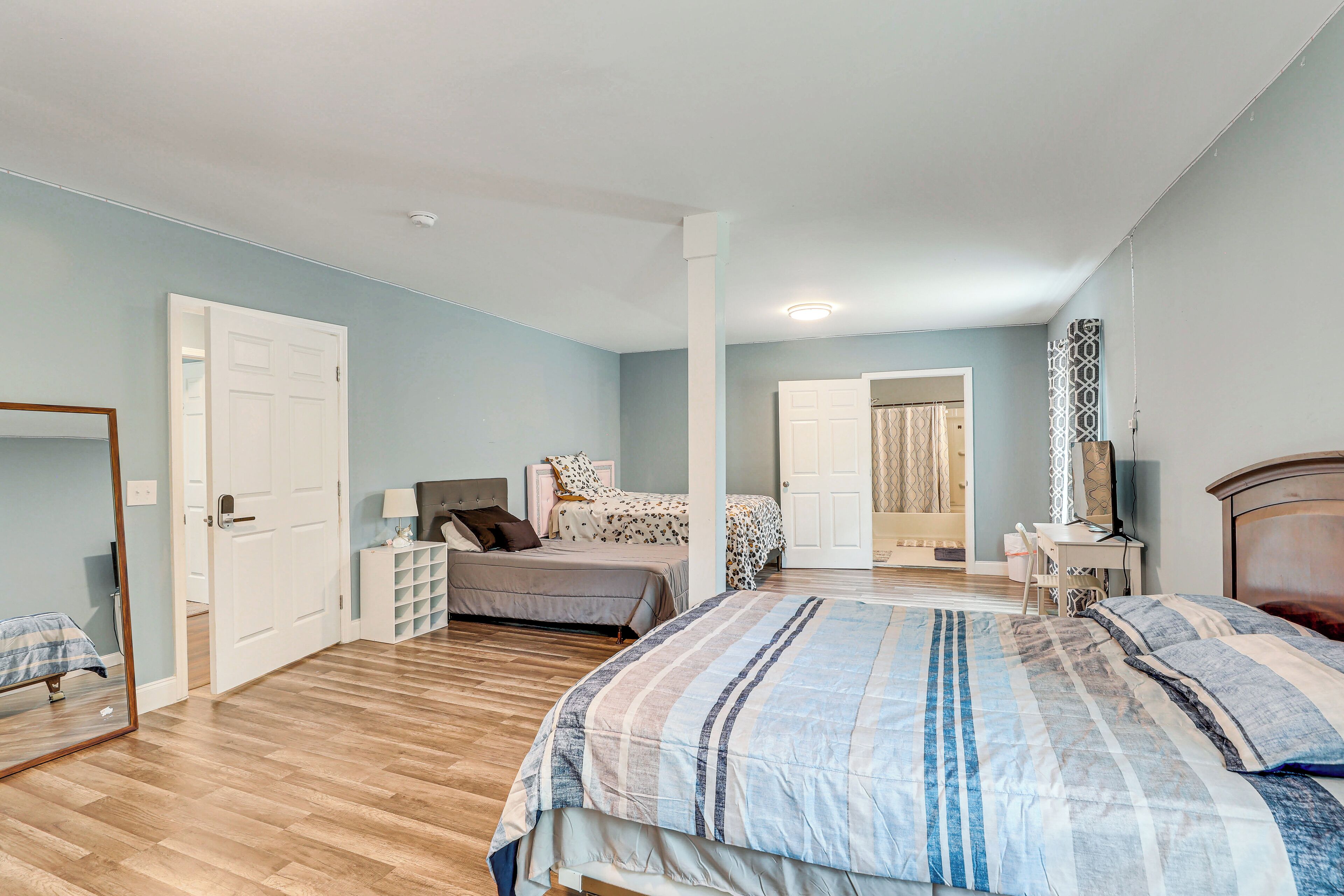 A bedroom with a large bed, a desk, and a chair. There is a door leading to another room.