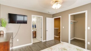 A bedroom with a bed, a TV mounted on the wall, a ceiling fan, and a hallway leading to a bathroom.