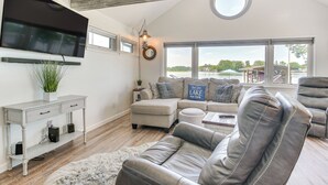 A living room with a sectional sofa, a flat-screen TV mounted on the wall, a round window, and a wooden ceiling beam. - Waterfront w/ Boat Dock: Retreat on Indian Lake (Belle Center)