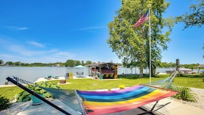 A hammock on a concrete patio with a view of a lake and a small building with an American flag. - Waterfront w/ Boat Dock: Retreat on Indian Lake (Belle Center)