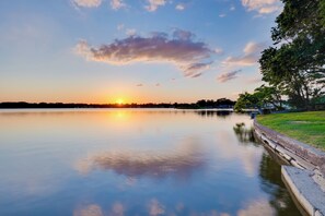 Un lago tranquilo al atardecer con cielo despejado, una orilla cubierta de hierba y una costa lejana.