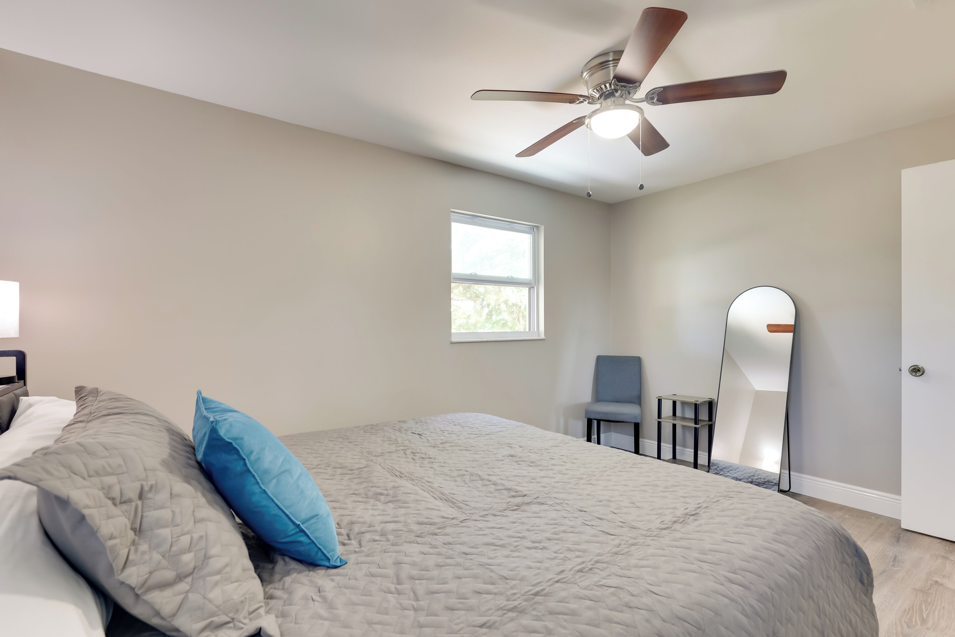 Un dormitorio con cama, ventilador de techo, ventana, silla y espejo.