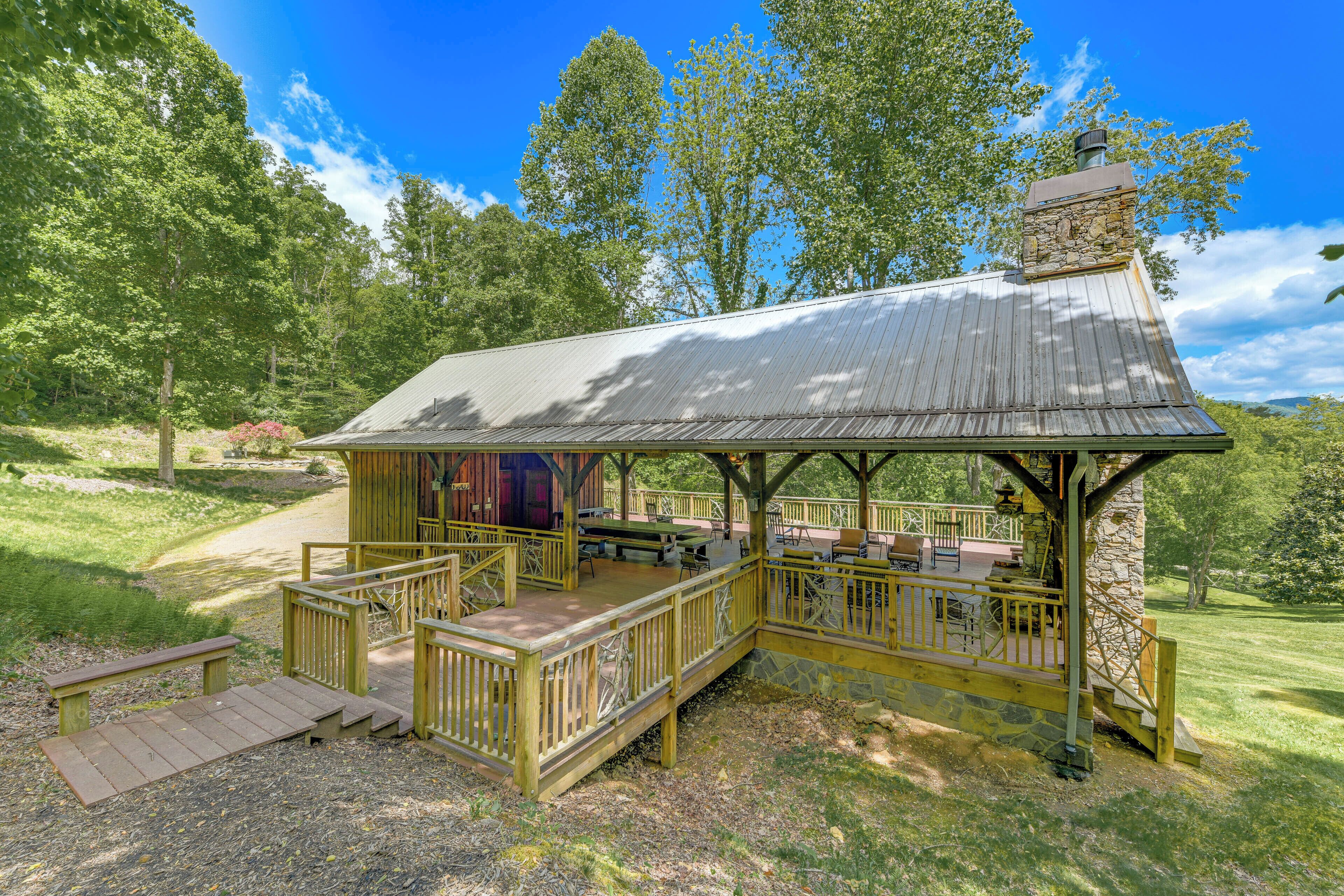 A rustic wooden pavilion with a stone chimney, surrounded by a deck and picnic tables, set in a lush green park.