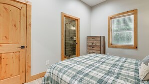 Un dormitorio con colcha a cuadros, armario de madera, cabina de ducha de cristal y ventana con persianas.