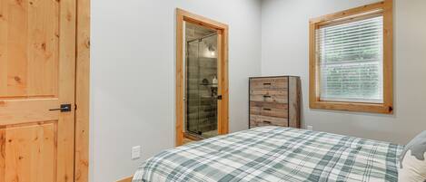 A bedroom with a plaid bedspread, a wooden wardrobe, a glass shower enclosure, and a window with blinds.