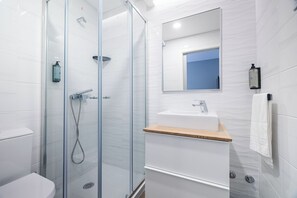 A modern bathroom with a glass-enclosed shower, a white sink with a wooden countertop, and a mirror above the sink. - Monturi Hostel (Ponta Delgada)