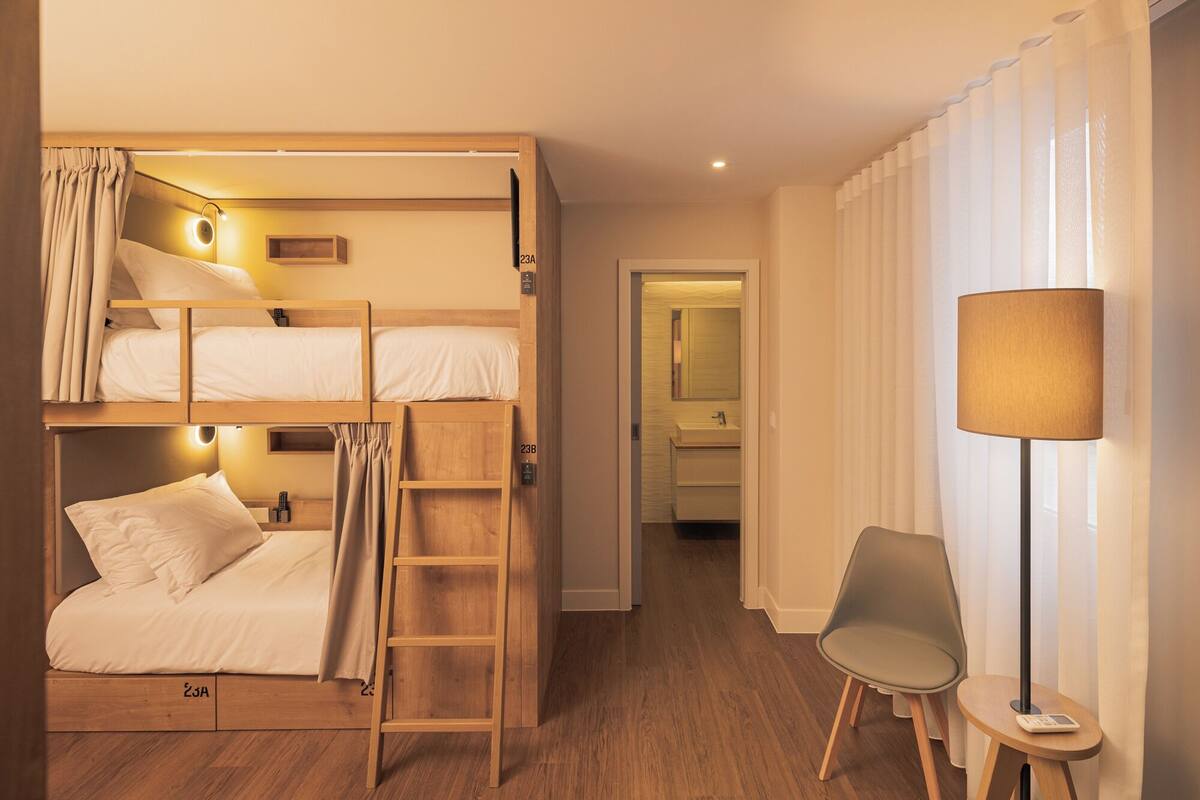A bunk bed room with a ladder, a chair, a lamp, and a bathroom visible in the background.