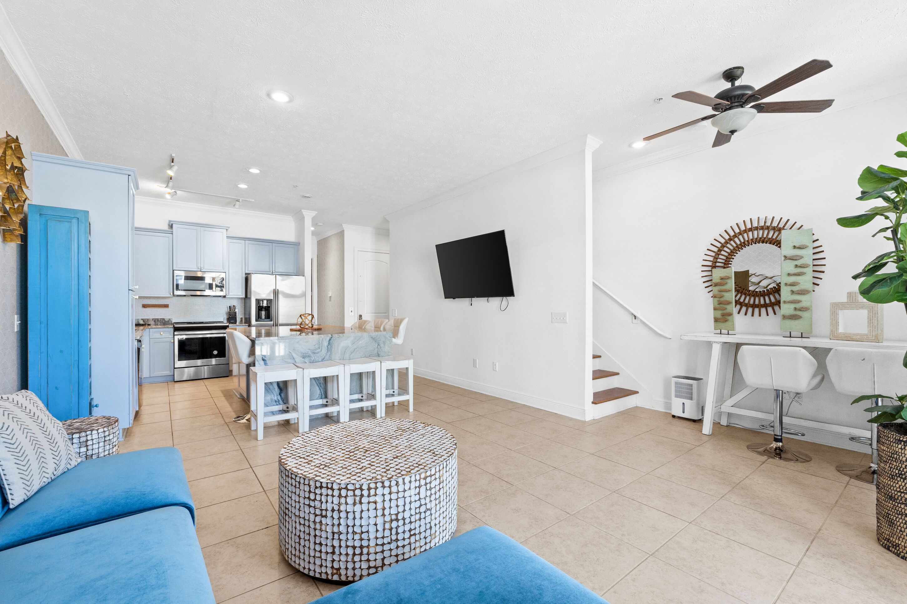 Una sala moderna con un sofá azul, un otomano estampado, una zona de comedor con una mesa blanca y una cocina al fondo.