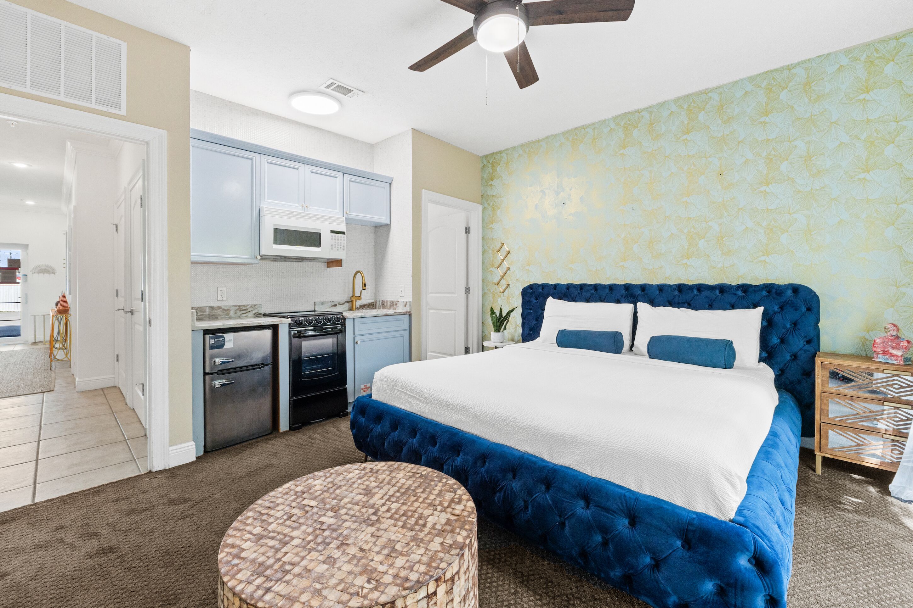 A bedroom with a large bed, a small kitchenette, and a ceiling fan.