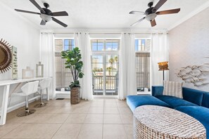 Un salon spacieux avec un canapé bleu, un bureau blanc avec deux chaises, une grande plante en pot et un balcon offrant une vue sur les immeubles et les palmiers.