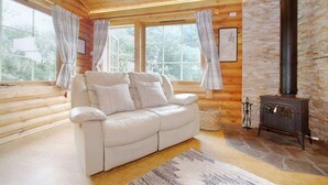 Interior acogedor de una cabaña de troncos con un sofá de cuero blanco, ventanas amplias, una chimenea de piedra y un techo de madera rústica.
