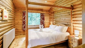 Ein Schlafzimmer in einem Blockhaus mit einem Himmelbett, Holzwänden, einem Fenster mit Vorhängen und einem Nachttisch mit Lampe.