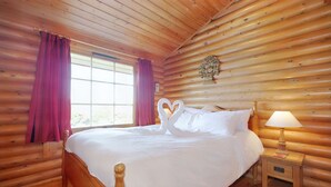 A wooden cabin bedroom with a bed, a nightstand with a lamp, and a window with curtains.
