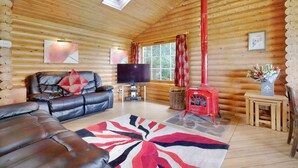 A log cabin interior with a leather sofa, a television, a red wood stove, and a large window with curtains.