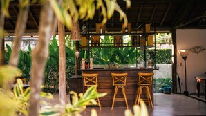 Zona de bar con taburetes de madera, estantes con botellas y vistas a un follaje exuberante.
