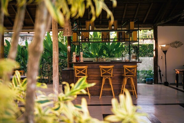 A bar area with wooden stools, shelves with bottles, and a view of lush greenery outside. - Ayla Arraial Pousada De Charme (Porto Seguro)