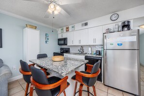 A compact kitchen with a dining table, chairs, a refrigerator, microwave, and a clock on the wall.