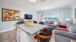 A modern living space with a kitchen island, dining area, and seating by a large window overlooking the sea. - Sunset Sands Retreat #4-Beachfront–Sleeps 4 (Fort Walton)