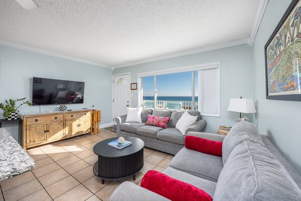 A living room with a sofa, a coffee table, a TV, and a view of the ocean.