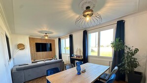 A modern living room with a wooden dining table, blue chairs, a grey sofa, and a large window with curtains. - CAP PLAGE 10 minutes' walk  (AUDIERNE)