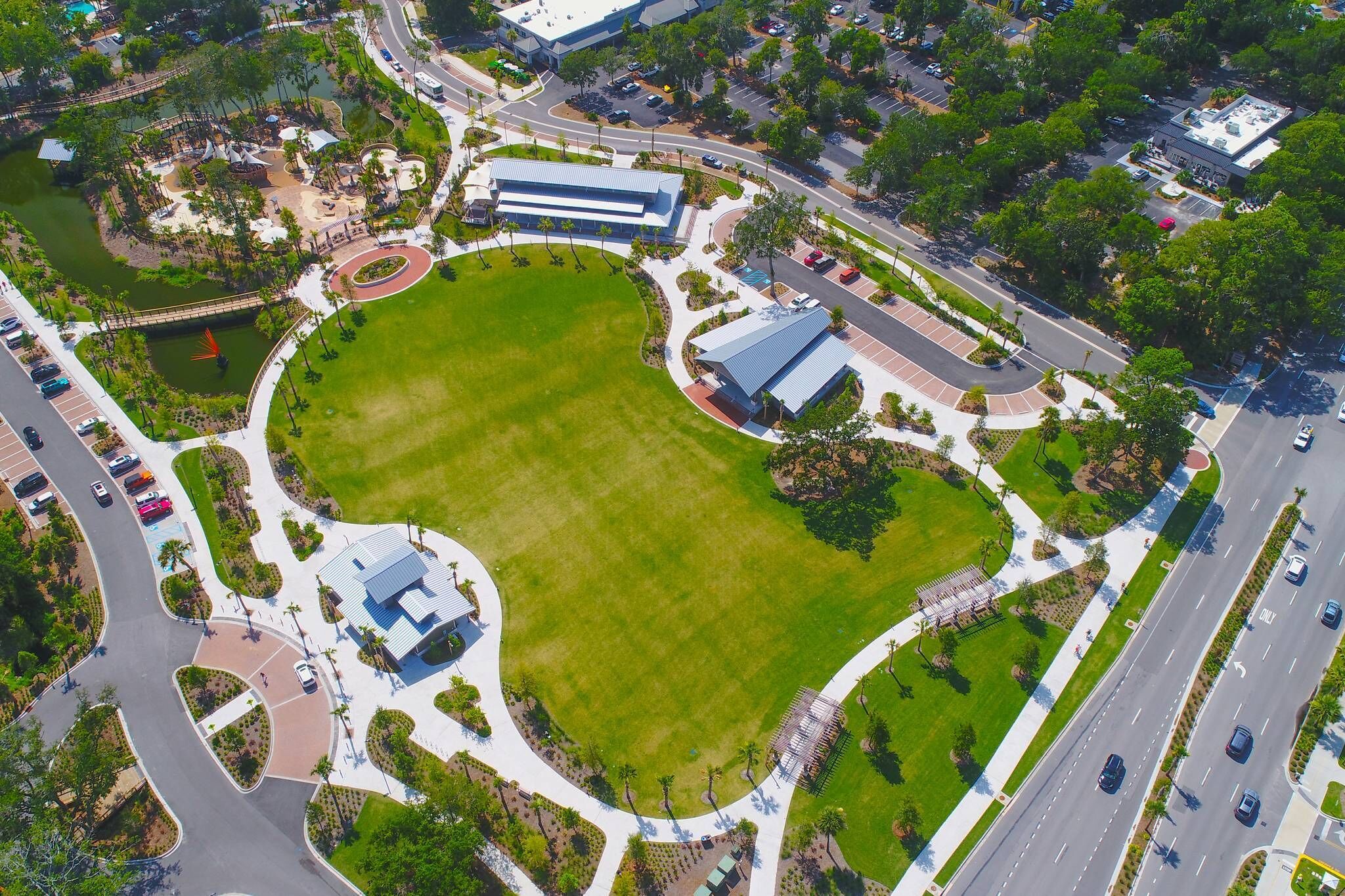 An aerial view of a park with a large green lawn, a building with a blue roof, and a parking area with cars.