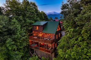 A two-story log cabin with a green roof, surrounded by dense forest, featuring a balcony with string lights and a visible deck area. - Prime Location w/ Views! Full Game Rm & Movie Rm! (Sevierville)