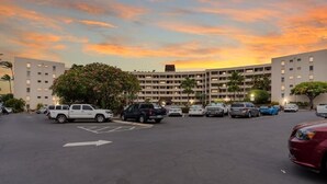A parking lot with multiple cars, a large building with a flat roof, and a tree in the foreground. - Royal Mauian, #511 2 Bedroom Condo by RedAwning (Kihei)